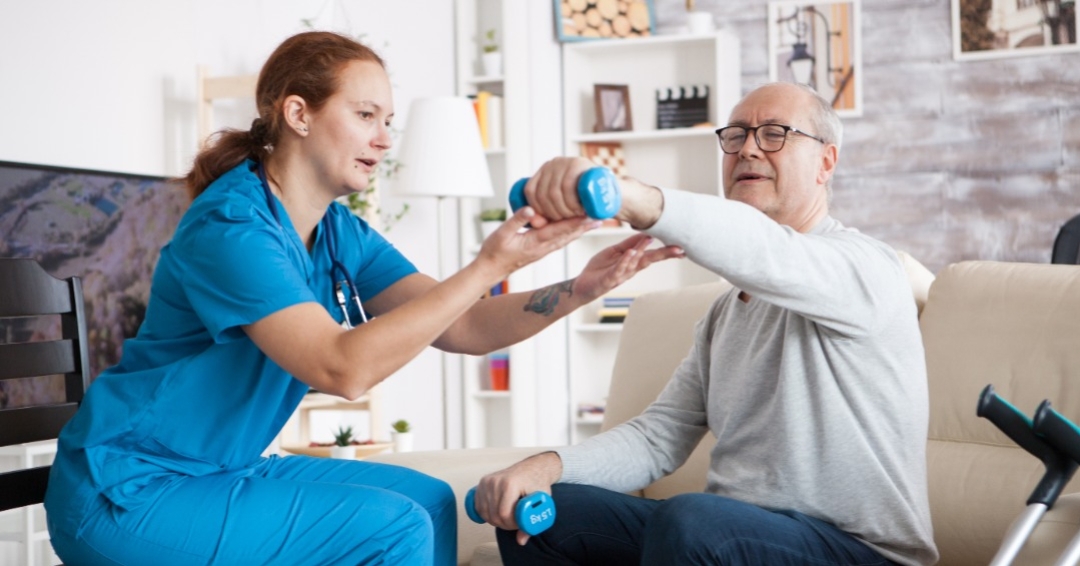Licensed physical therapist assisting an elderly man with strength training exercises using weights in a home setting